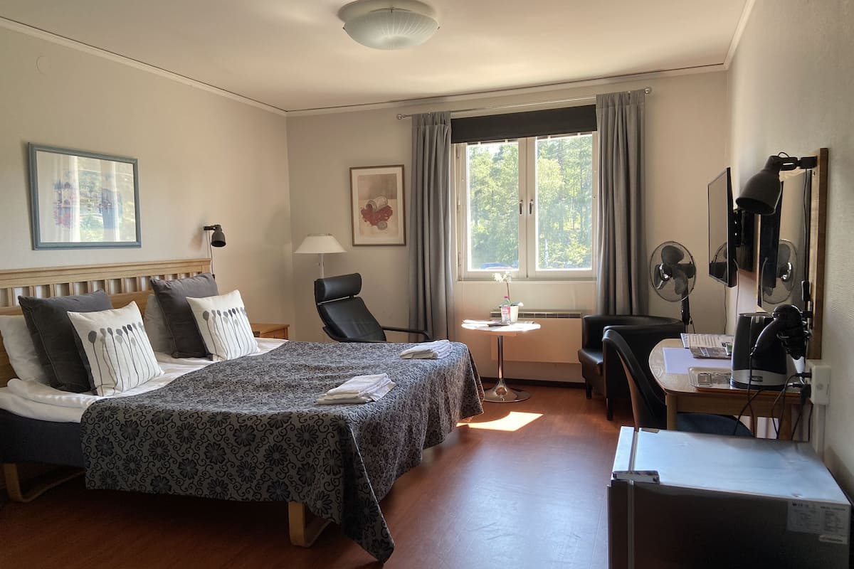 Une chambre d’hôtel comprenant un lit, un bureau, une chaise, une table, une lampe, un ventilateur et une fenêtre avec des rideaux.