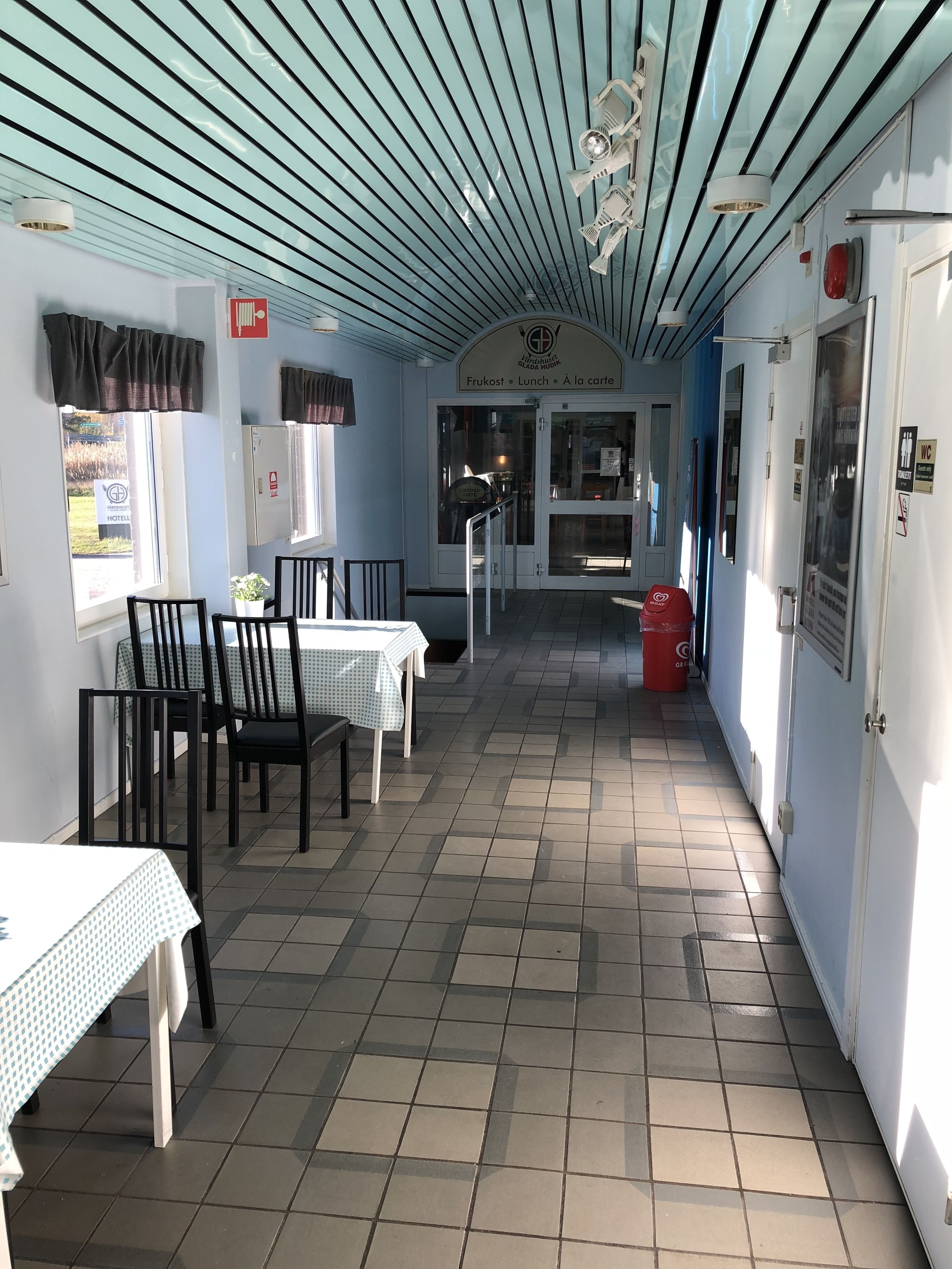 Un corridoio con tavoli e sedie, un pavimento piastrellato e una porta che dà su una stanza con il cartello che indica 'Frukost Lunch à la carte'.