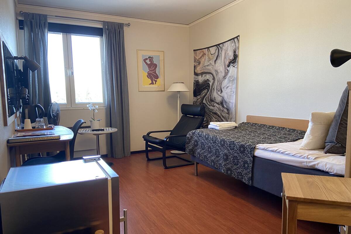 Une chambre d’hôtel comprenant un lit, un bureau, une chaise, une fenêtre avec des rideaux, un tableau au mur et une lampe.