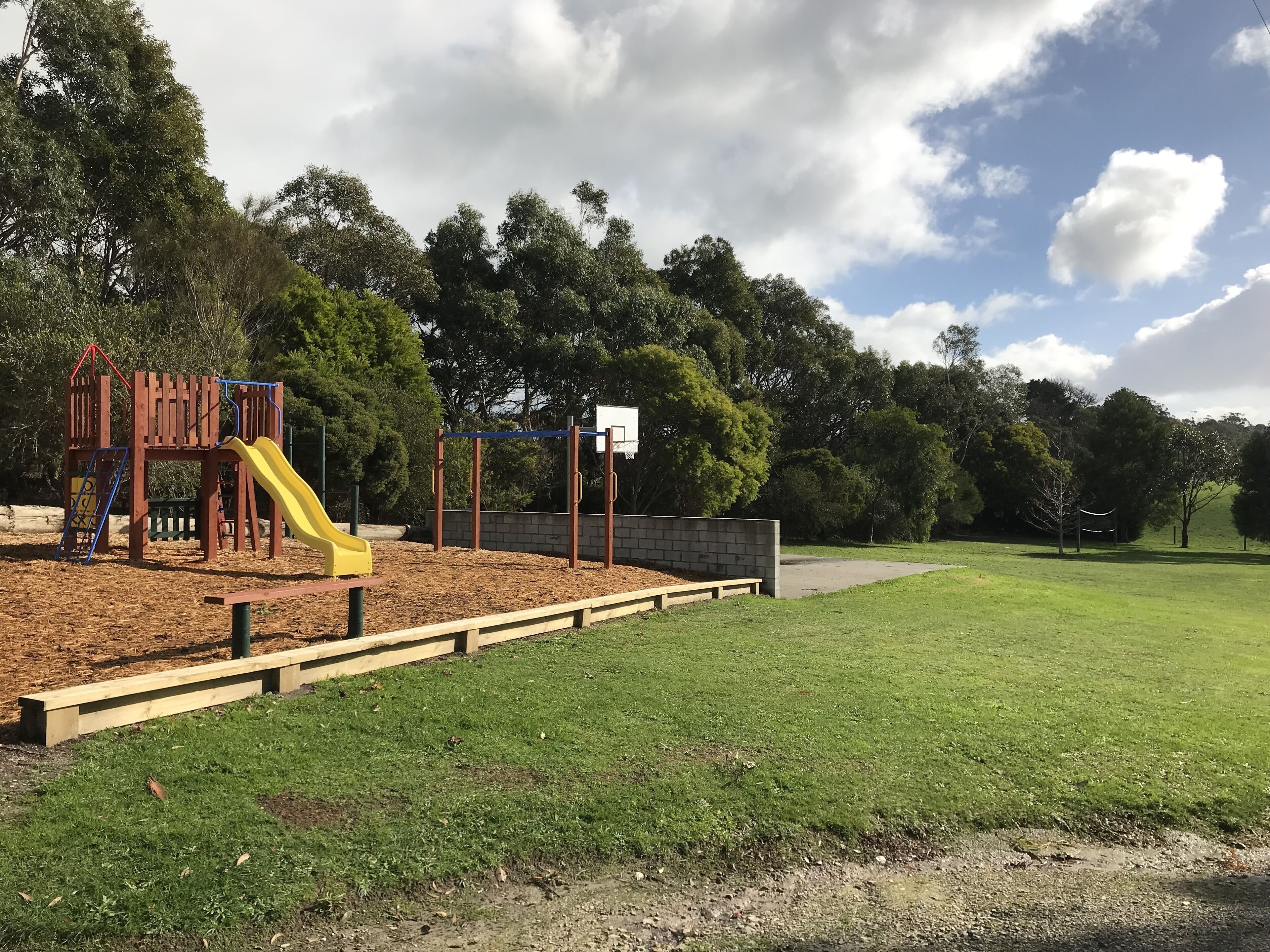 A playground with a slide and climbing structures, surrounded by a grassy area and trees.