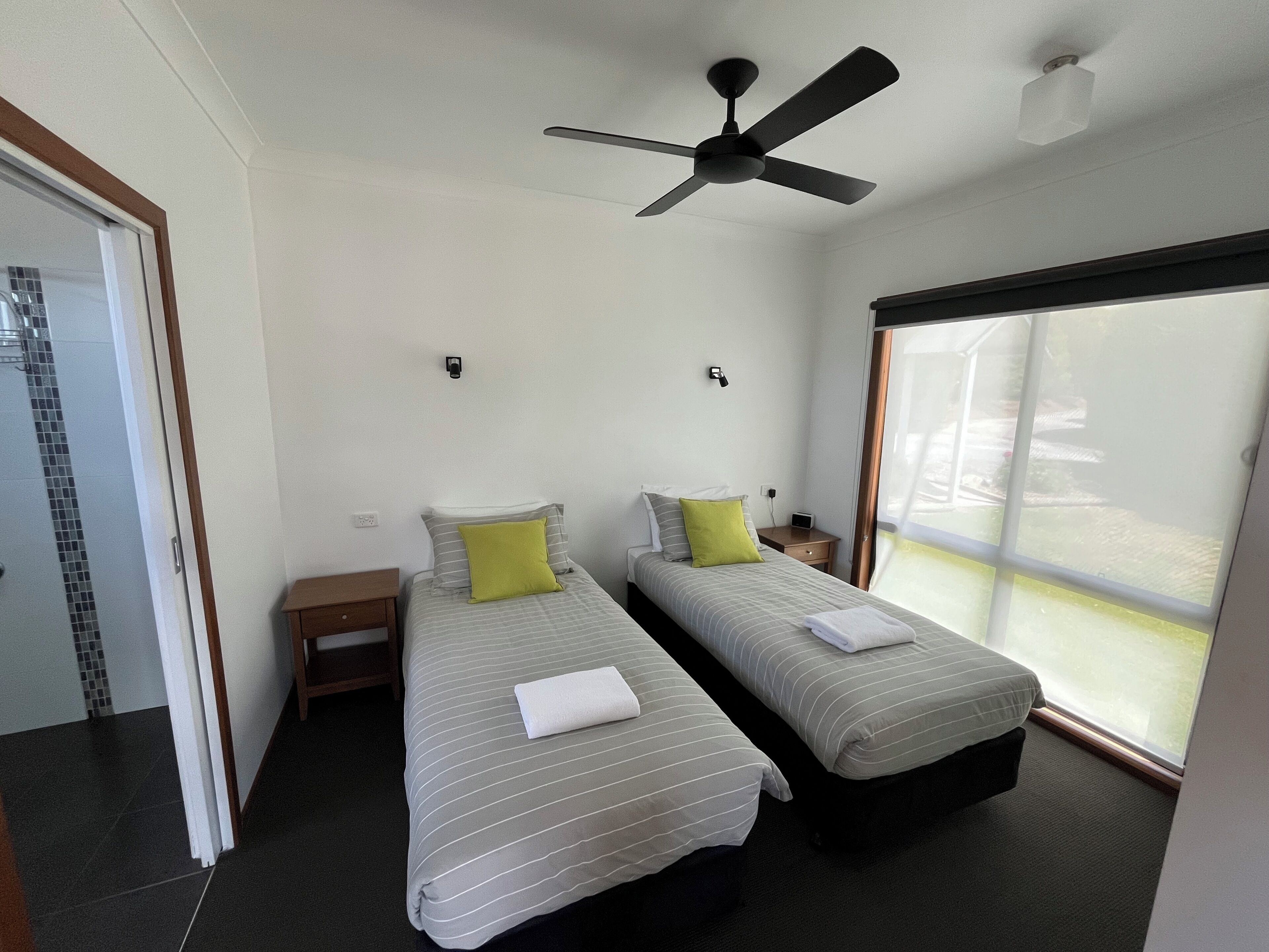 Una camera d'albergo con due letti singoli, una ventola a soffitto, una finestra con vista e un comodino con lampada.