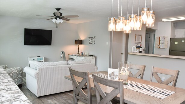 A modern living room with a sofa, dining table, and chairs. There is a ceiling fan, a TV mounted on the wall, and a kitchen area visible in the background. - Beachy Getaway–Cozy, Clean & Equipped w/ All Essentials! Relax, Enjoy & Refresh! (St Augustine Beach)