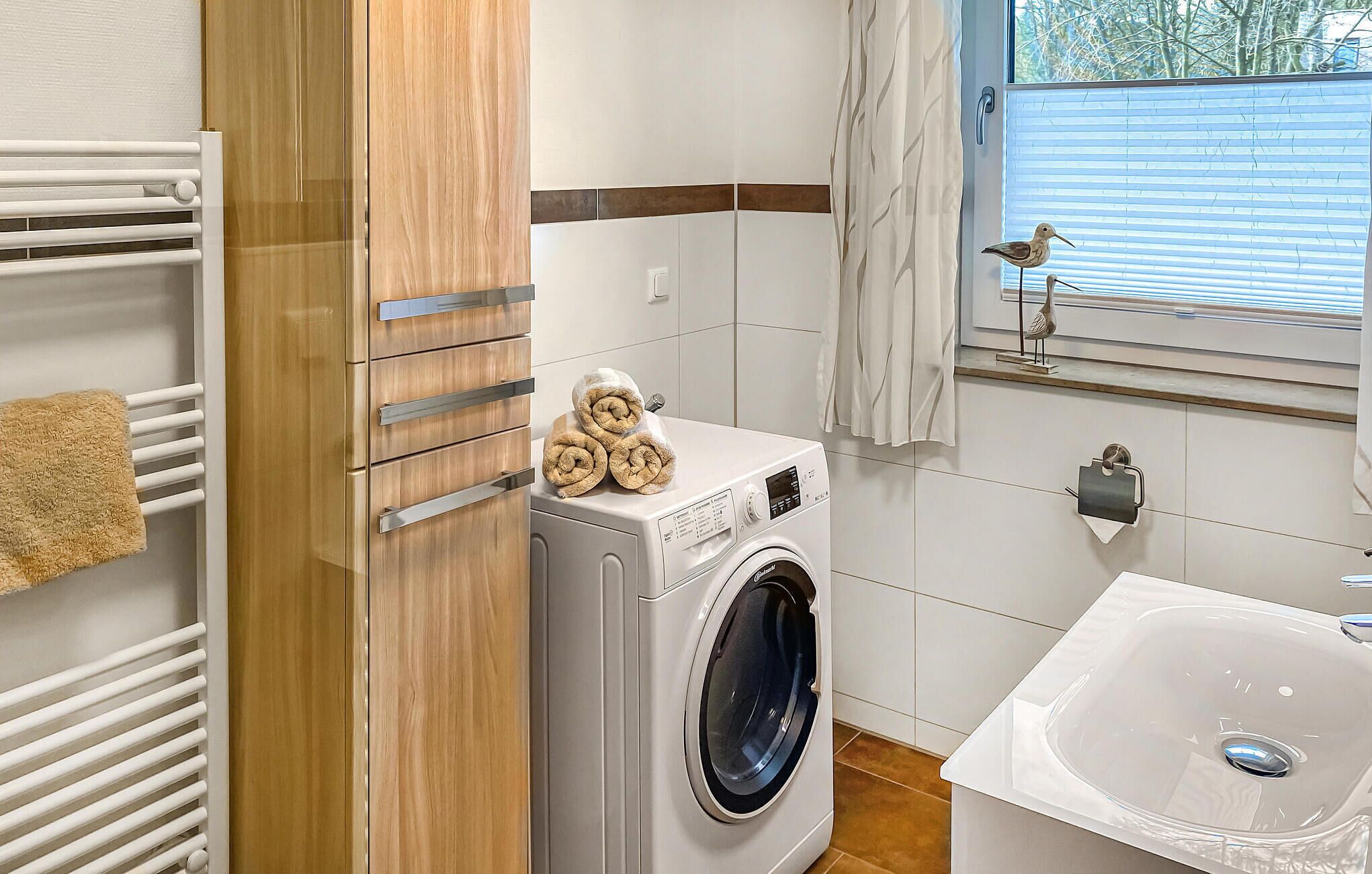 A compact laundry area with a front-loading washing machine, a drying rack, and a window with a view of trees.