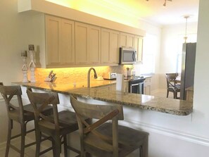 Una cocina con encimera de granito, sillas de madera, microondas y un lavamanos.