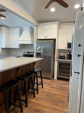 A modern kitchen with white cabinets, a stainless steel refrigerator, a microwave, and a built-in oven. There are two black bar stools and a ceiling fan. - 4 Bed 3.5 Bath Pool Mid Island, Quiet Garden lined Street, steps from the Beach! (Ocean Isle Beach)