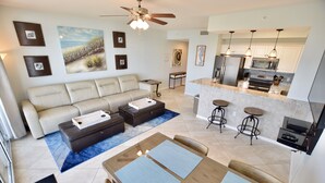 A modern living room with a sofa, coffee table, and dining area. There is a kitchen with a refrigerator, microwave, and bar stools. - Destin West Gulfside #603 ~WOW! ~Spectacular Penthouse View! ~Free Beach Service (Fort Walton Beach)