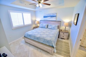 A bedroom with a bed, two bedside tables, a ceiling fan, a window with blinds, and a beach-themed painting on the wall.