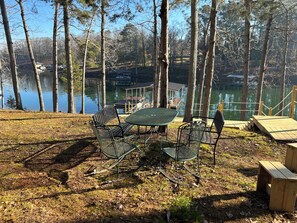 A lakeside outdoor seating area with metal chairs and a table, surrounded by trees and a dock extending into the water.