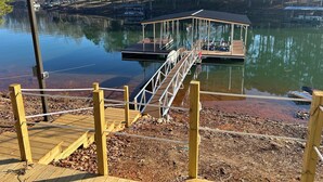 Un muelle de madera que conduce a un área de estar cubierta junto a un lago tranquilo, rodeado de árboles.