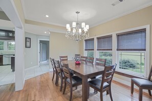 A dining room with a large wooden table, six chairs, a chandelier, and large windows with blinds.