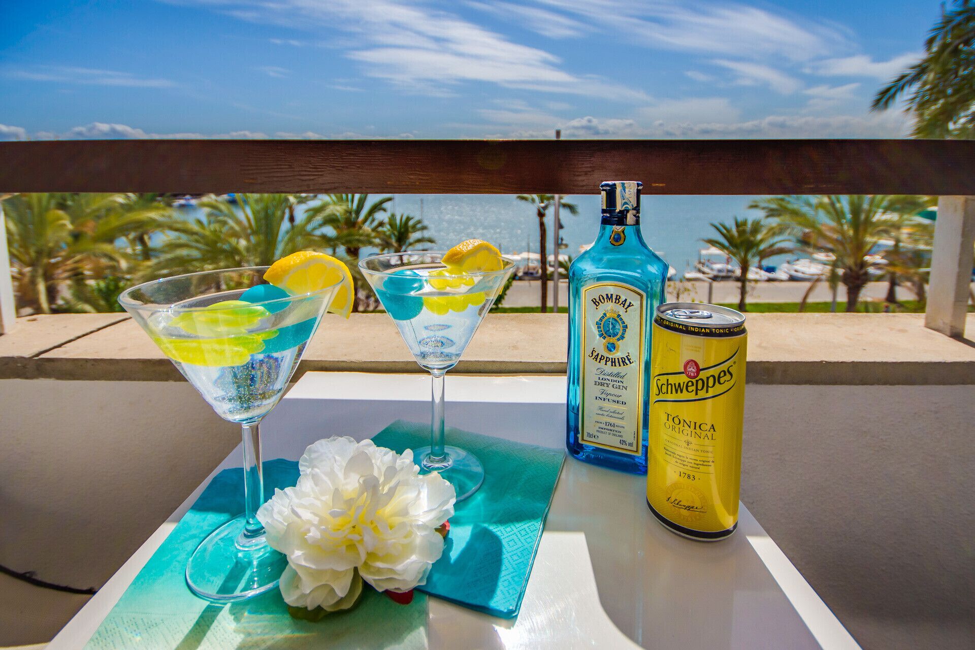 A table with two martini glasses, a bottle of Bombay Sapphire gin, a can of Schweppes tonic water, and a white flower on a blue napkin.