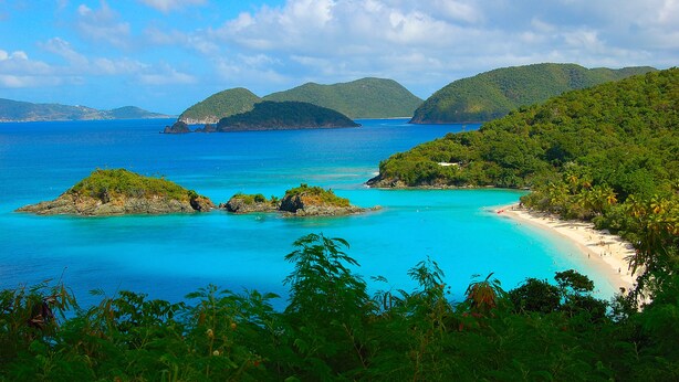 A coastal landscape with clear turquoise waters, green hills, and a sandy beach.