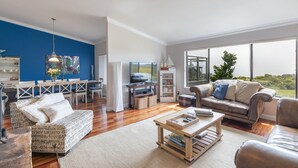 Uma sala de estar espaçosa com uma parede azul de destaque, uma janela ampla com vista, um espaço de estar acolhedor com sofá e poltronas, e uma área de jantar com mesa e cadeiras.