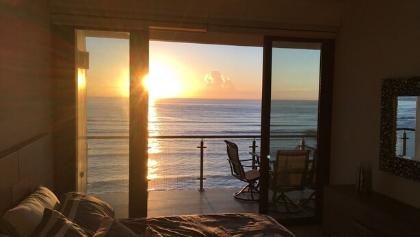 A bedroom with a large sliding glass door leading to a balcony with a view of the ocean at sunset. - Luxury Penthouse Beachfront Condo with Private Rooftop (Akumal)