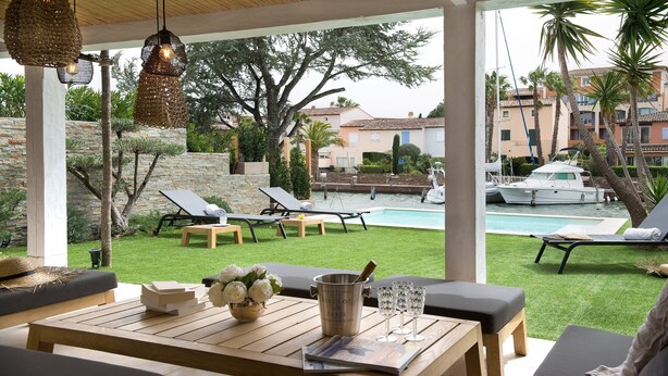 Un espace aménagé au bord de la piscine, avec des chaises longues, une table en bois ornée d’un vase de fleurs et une vue sur un port de plaisance rempli de bateaux.
