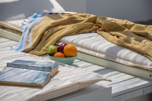 A bed with a white mattress, a blue and white striped blanket, and a bowl of fruit including apples, oranges, and a banana. - The Veranda of Gavrion - Premium Cycladic House (Andros)