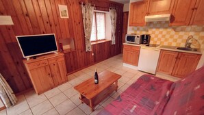 Une pièce aux murs lambrés, avec un téléviseur, une table en bois sur laquelle se trouve une bouteille, un canapé rouge et un coin cuisine équipé d’un four à micro-ondes et d’une machine à café.