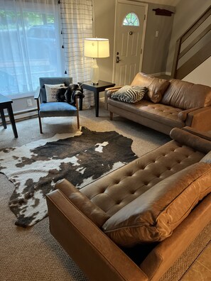 Una sala de estar con un sofá de cuero marrón, un sillón azul, una mesita con una lámpara y un tapete de piel de vaca.