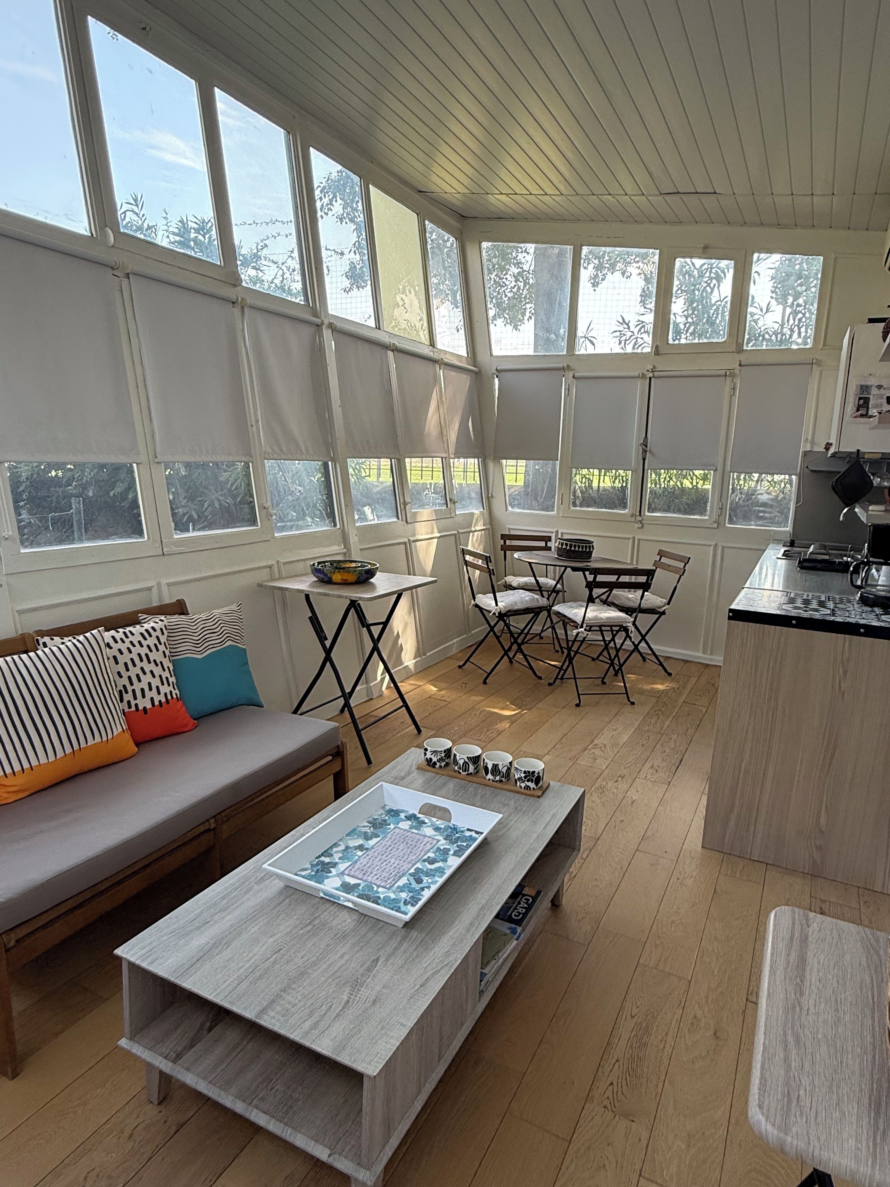 A sunlit room with a wooden floor, a sofa with patterned cushions, a coffee table, and a dining area with chairs and a table.