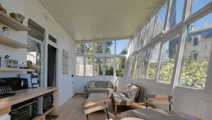 A bright, sunlit room with a kitchen area, a seating area with a sofa and chairs, and large windows offering a view of trees and buildings.