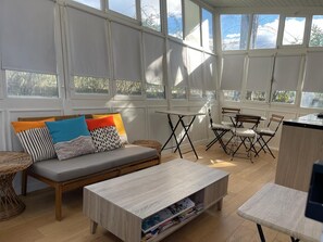 A bright, sunlit room with a sofa, a wooden coffee table, and a dining area with chairs and a table. - Charming air-conditioned cottage "the cabin" and its private and independent garden (Sommières)