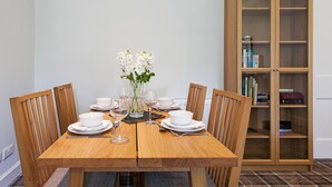 A dining table set with white bowls, plates, and wine glasses, flanked by wooden chairs. A wooden cabinet with glass doors is to the right. - The Old Manse House - Apartment (Ayr)