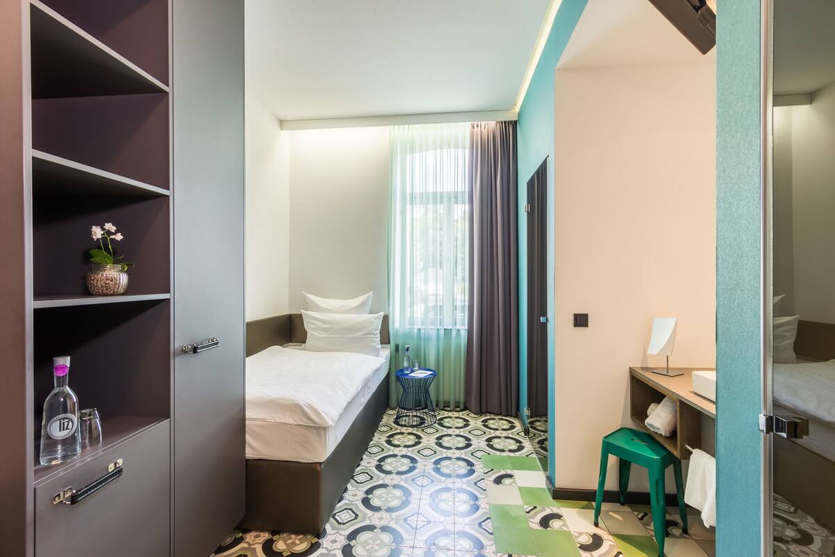 A hotel room with a bed, a small table, a chair, a shelf with a bottle, and a window with curtains.