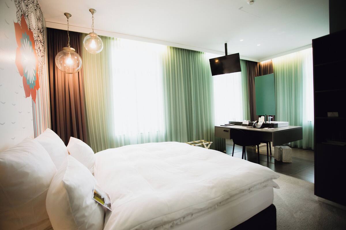 A modern hotel room with a large bed, a desk, a flat-screen TV, and a view of the city.
