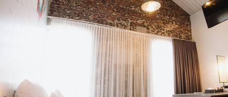 A bedroom with a brick wall, a bed with white linens, a desk with a lamp, and a large window with curtains.