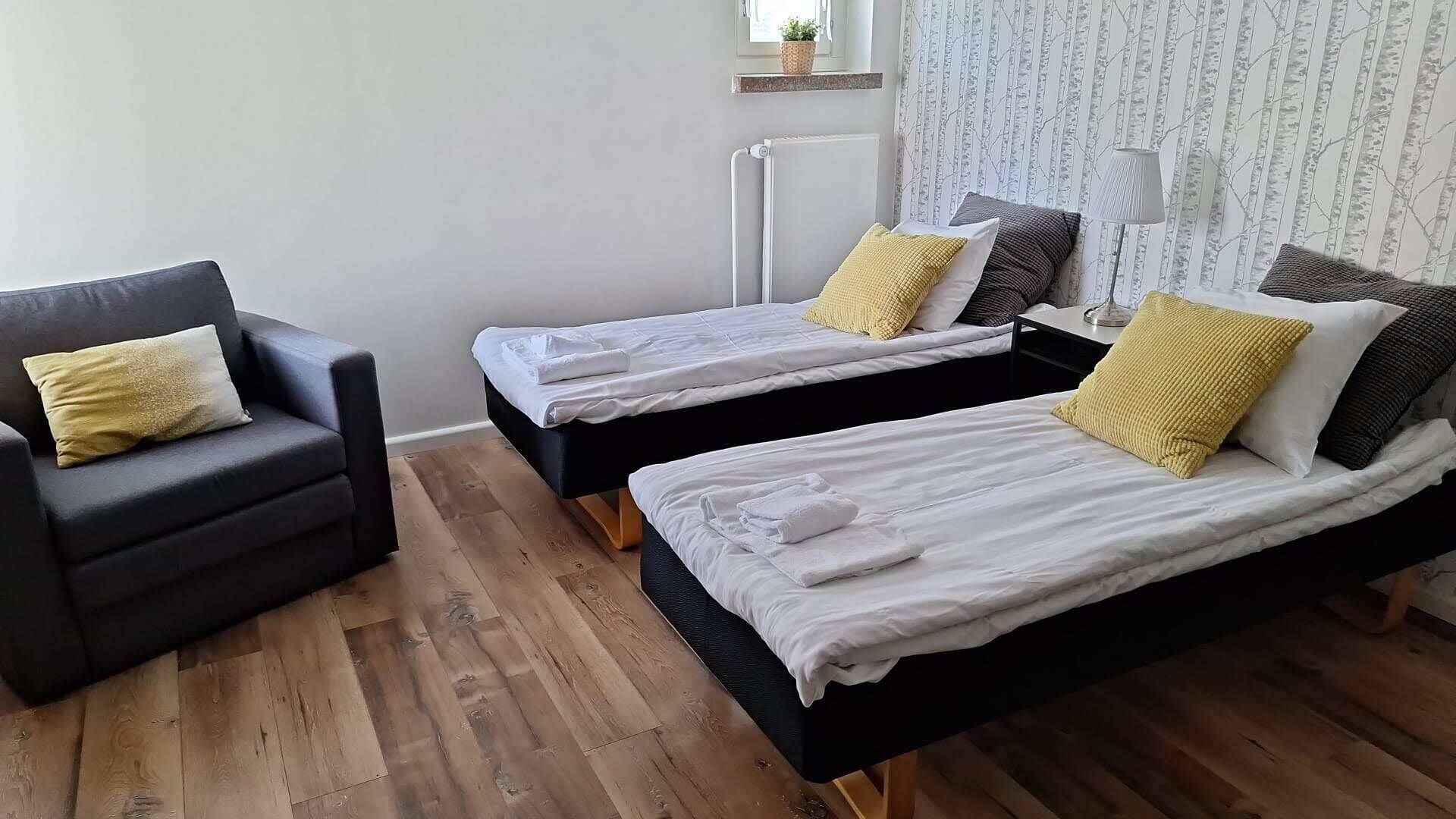 Une chambre avec deux lits simples, un fauteuil noir et une fenêtre donnant sur des arbres.