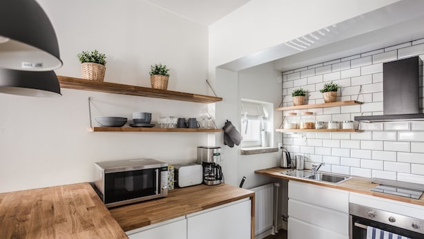 A modern kitchen with white cabinets, wooden countertops, and white tiled backsplash. There are shelves with plants and kitchen items, a microwave, toaster, and coffee maker. - Haave Apartments (Valkeakoski)