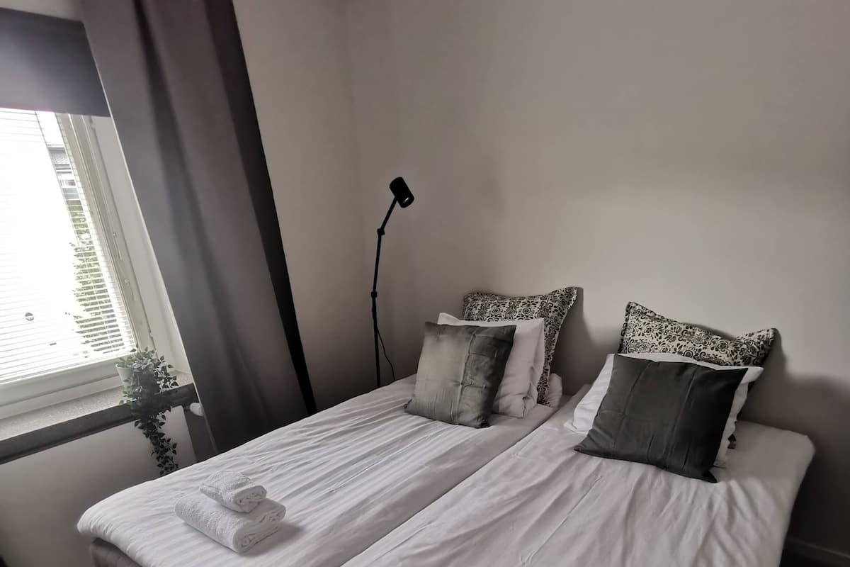 A neatly made bed with white linens and two pillows, a standing lamp, and a window with curtains.