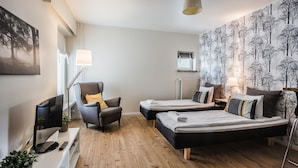 A modern hotel room with a wooden floor, a flat-screen TV, a desk with a lamp, two single beds, a chair, and a large window. - Haave Apartments (Valkeakoski)