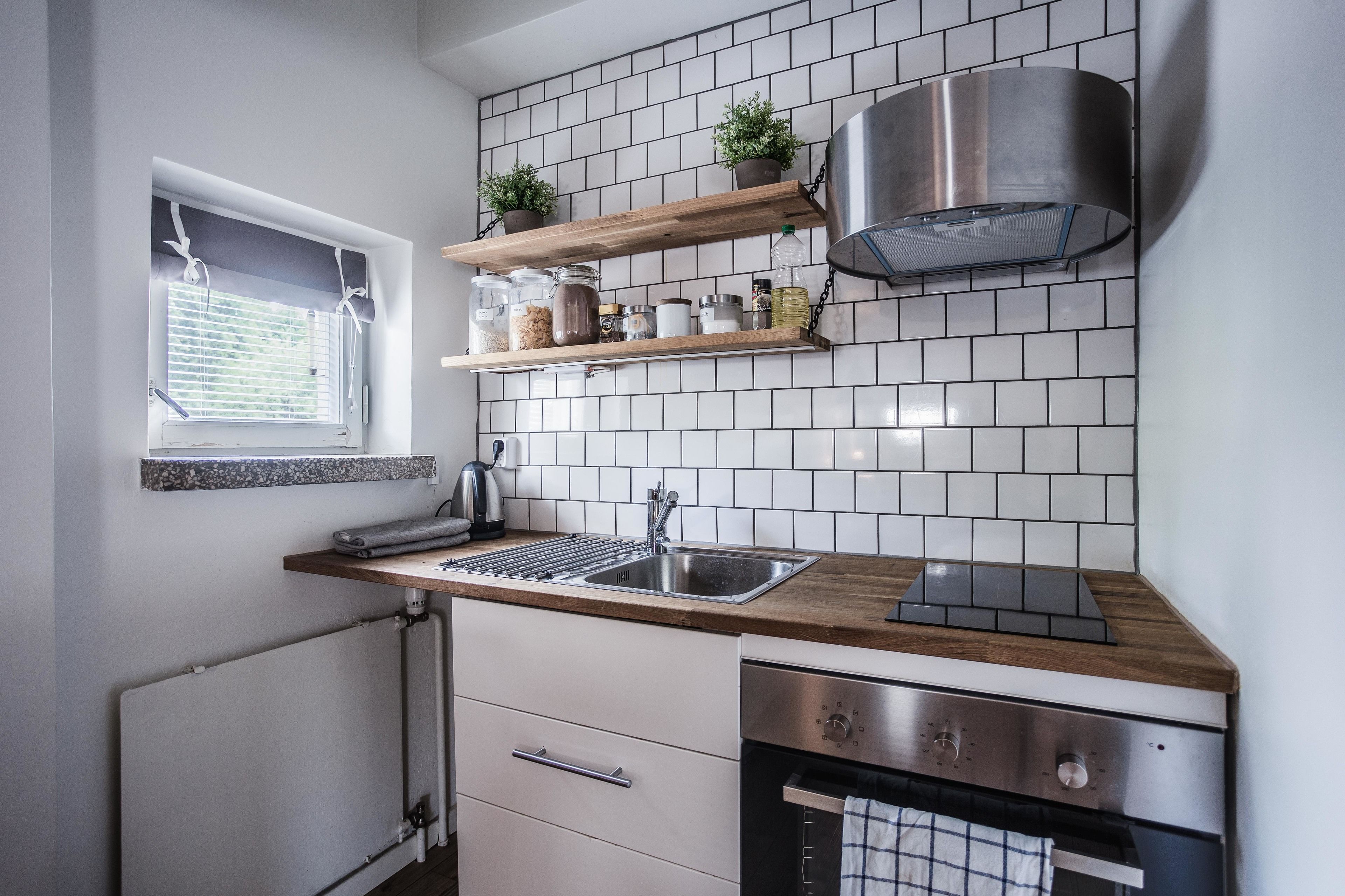 Une cuisine moderne avec des carreaux de céramique blancs disposés en motif métro, des étagères en bois, une hotte de cuisine en acier inoxydable et une fenêtre avec des stores.