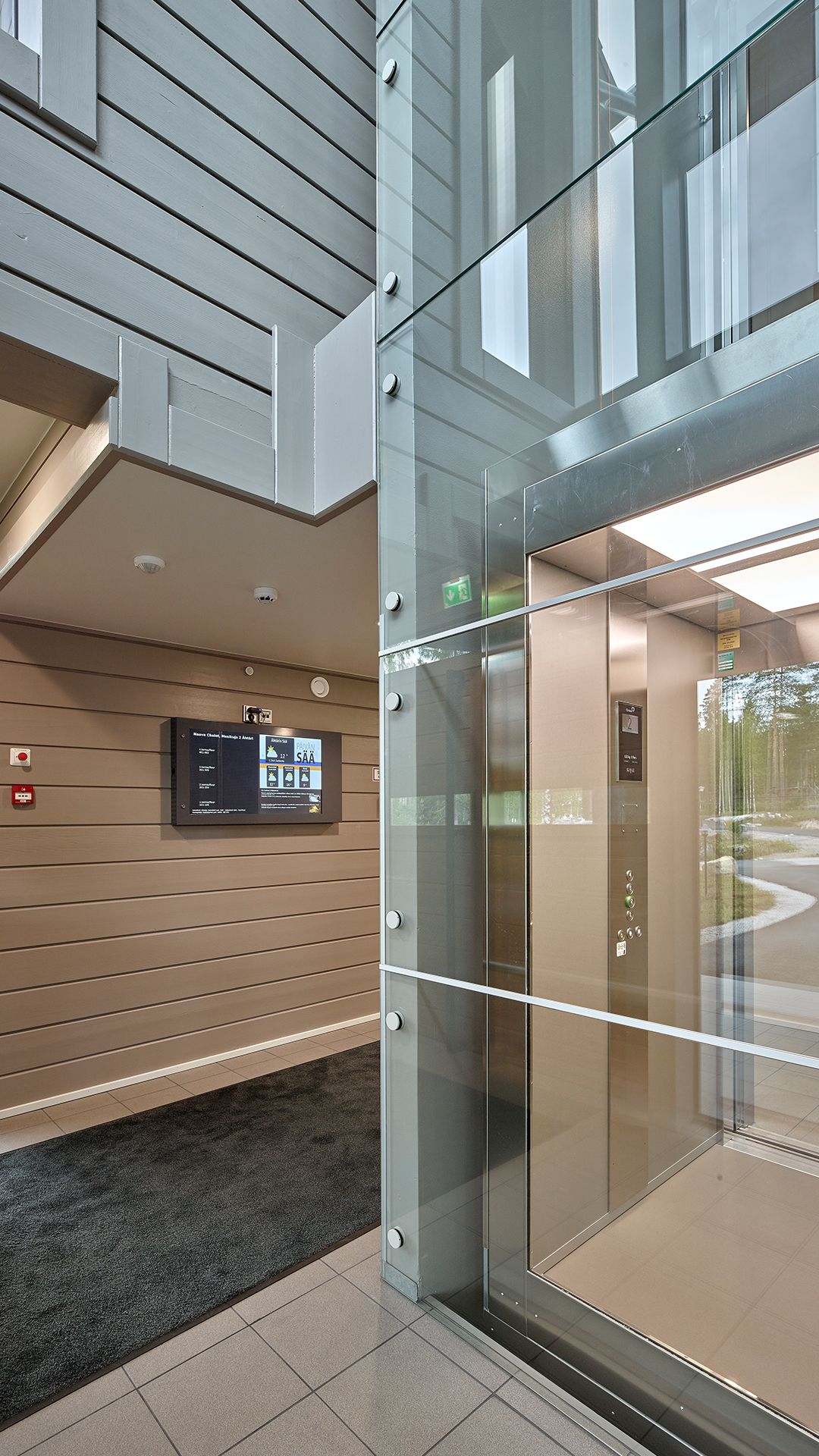 An elevator with a glass door, a digital information board, and a beige wall with horizontal lines.
