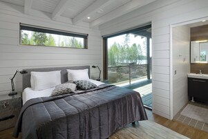 Quarto com cama, luminárias de cabeceira, mesa de cabeceira, janela com vista para árvores e varanda.