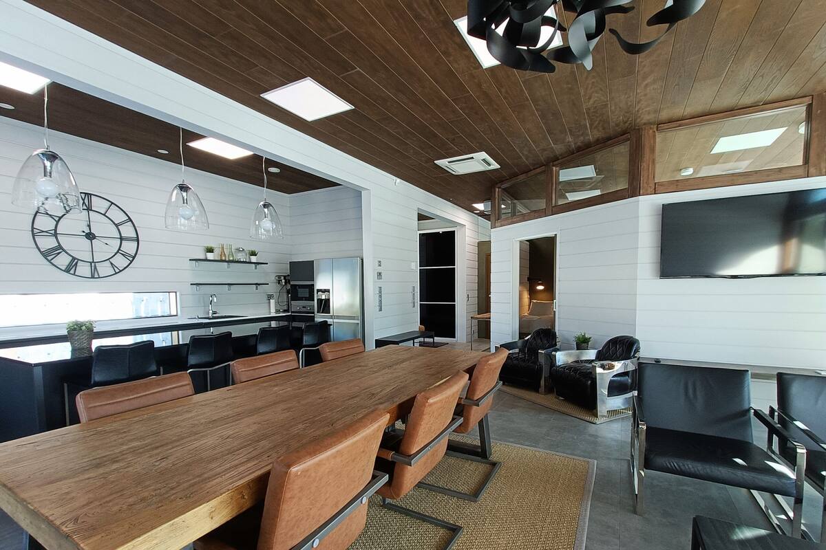 A modern kitchen with built-in appliances, a dining area with a long wooden table and leather chairs, and a large flat-screen TV mounted on the wall.