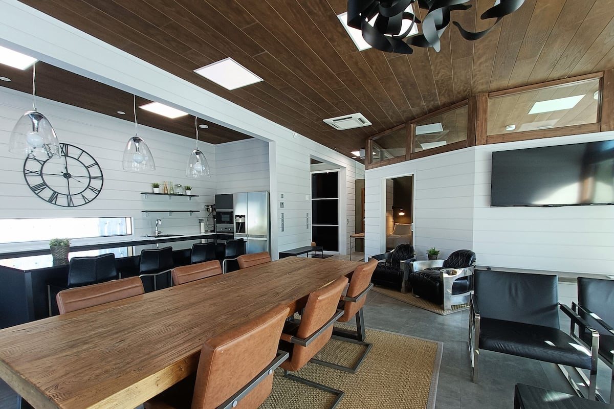 Una cocina moderna con electrodomésticos integrados, una zona de comedor con una mesa de madera alargada y sillas de cuero, y un televisor de pantalla plana grande montado en la pared.