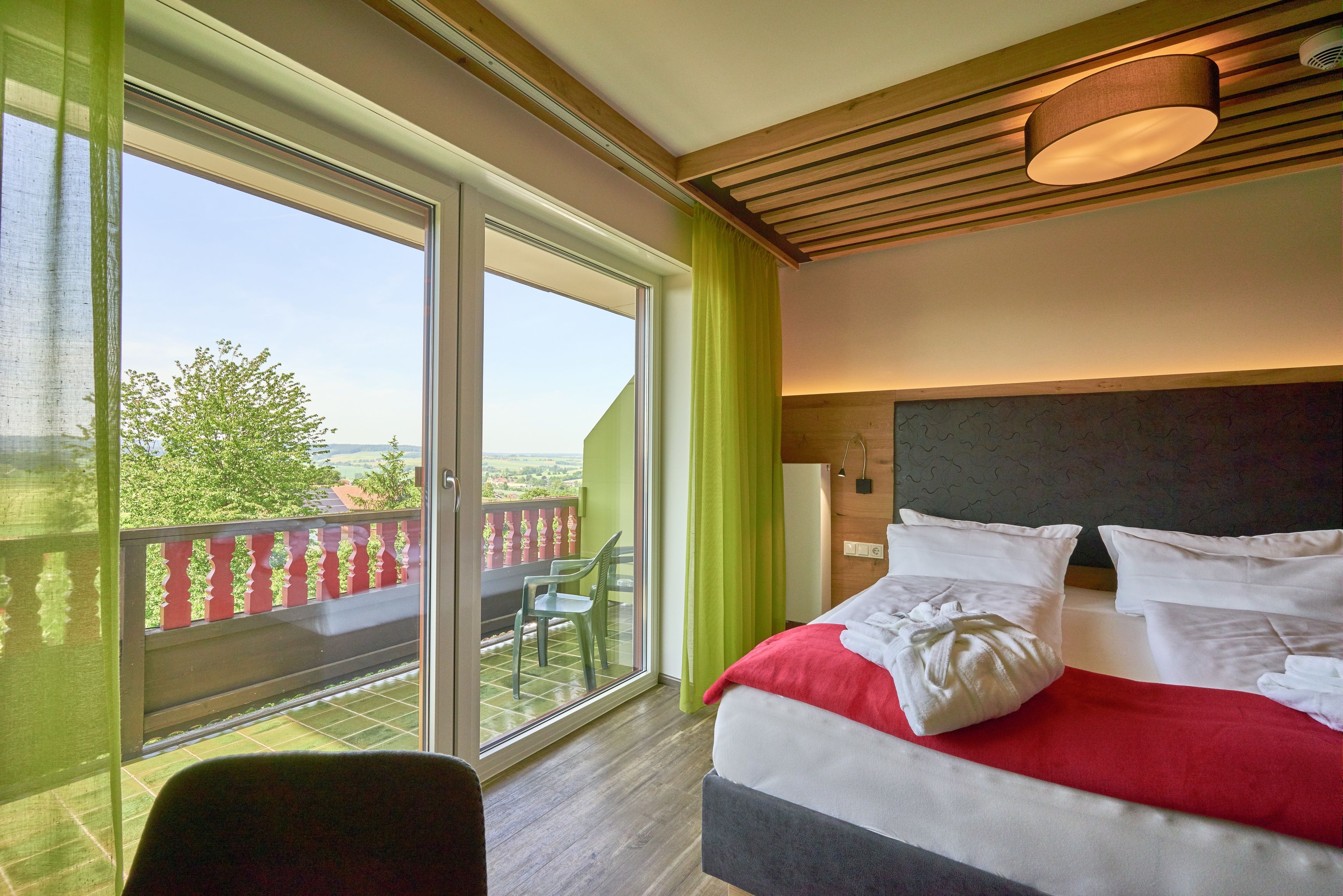 Quarto de hotel com uma grande porta de vidro deslizante que dá para uma varanda, cama com roupa de cama branca e vermelha, e teto de madeira.