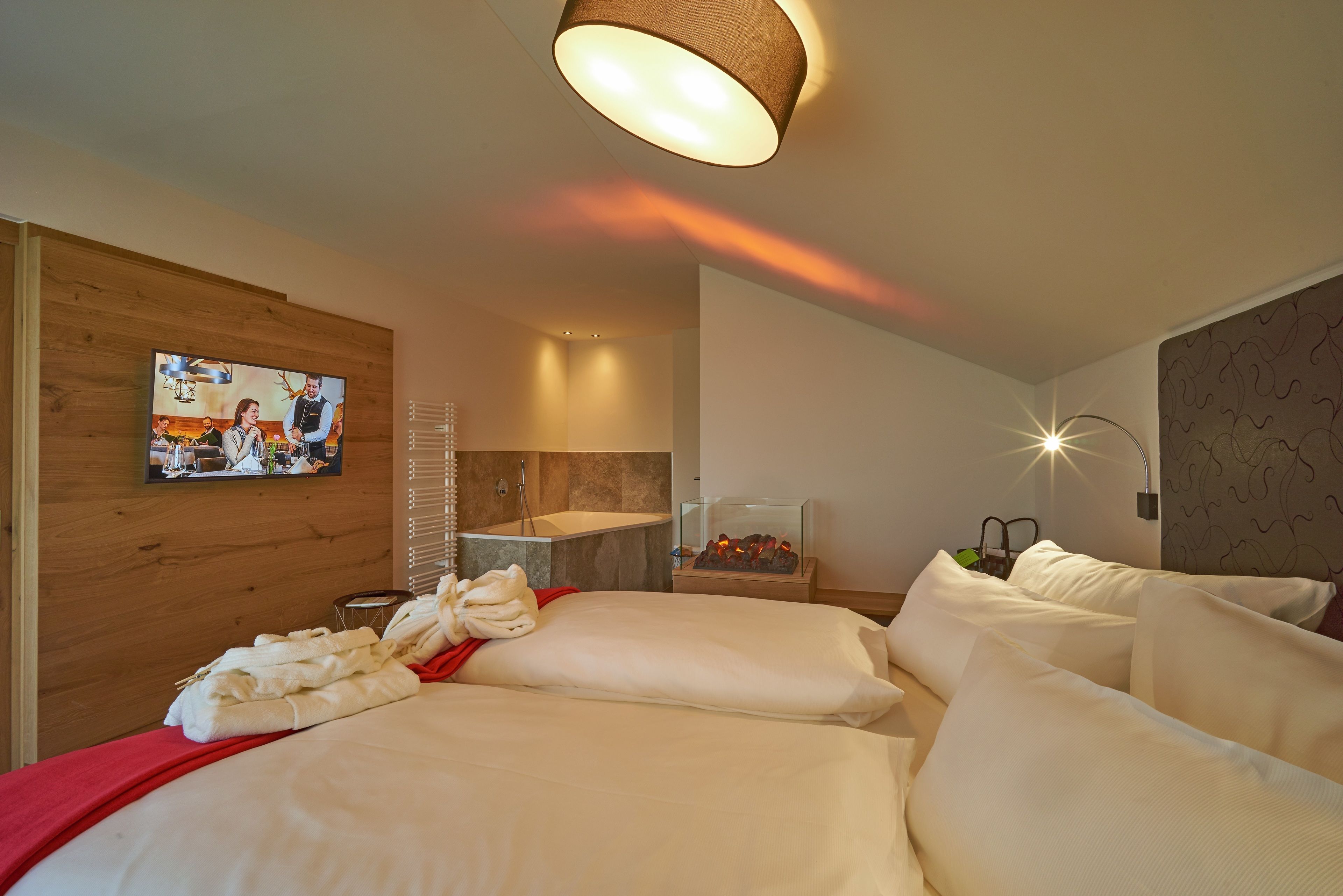 A hotel room with a large bed, a TV mounted on the wall, a bathroom with a bathtub, and a nightstand with a lamp.