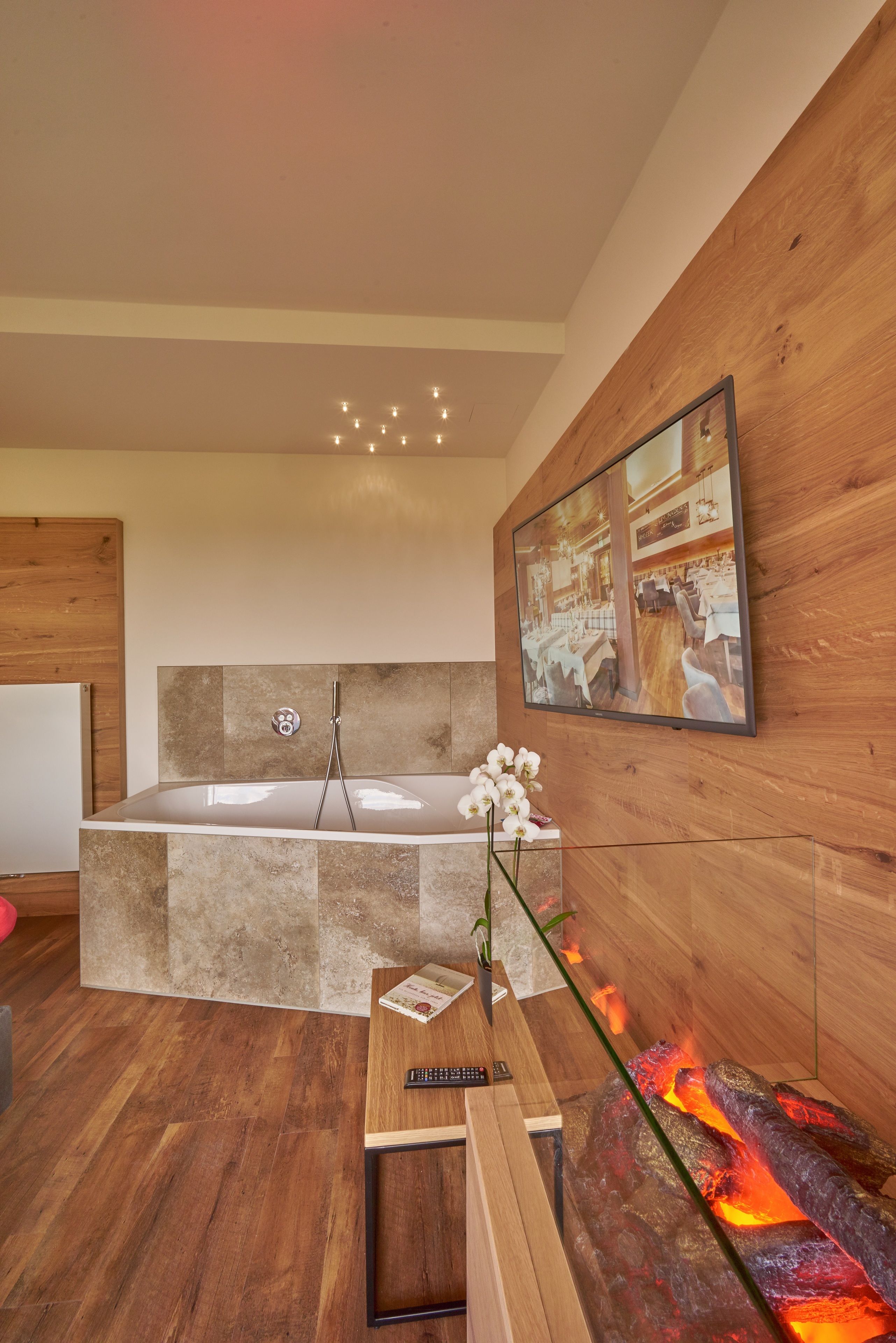 Une salle de bain moderne avec une grande baignoire, un mur en bois, un téléviseur fixé au mur et une table en verre avec une télécommande.