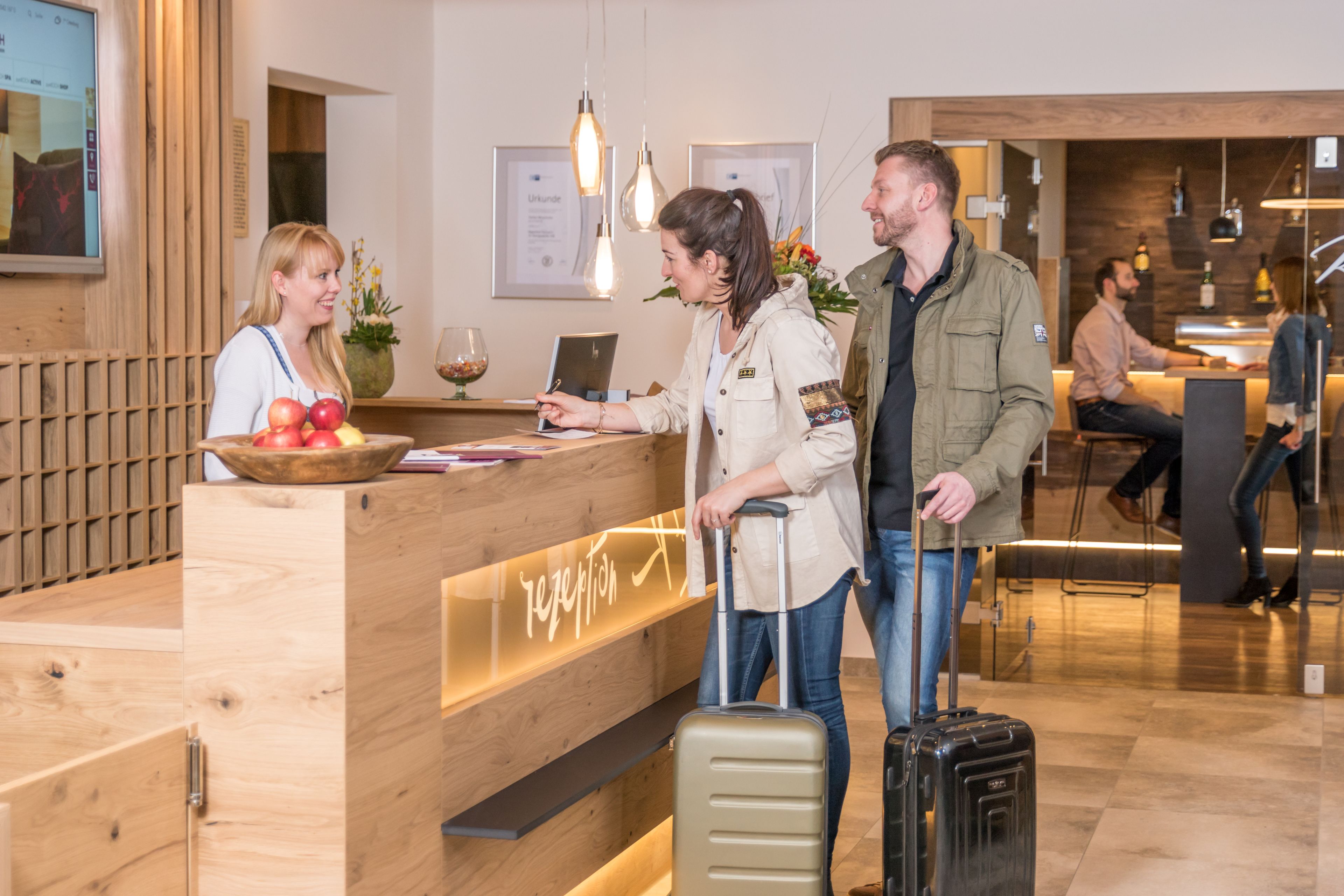 A reception desk with a wooden counter, a laptop, and a fruit bowl. There are two people standing in front of the desk, one with a suitcase.