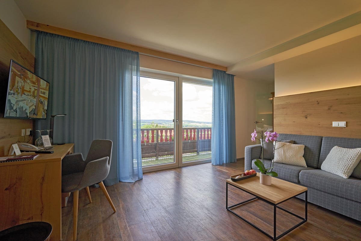 A modern living room with a wooden desk, a grey sofa, and a balcony with blue curtains.