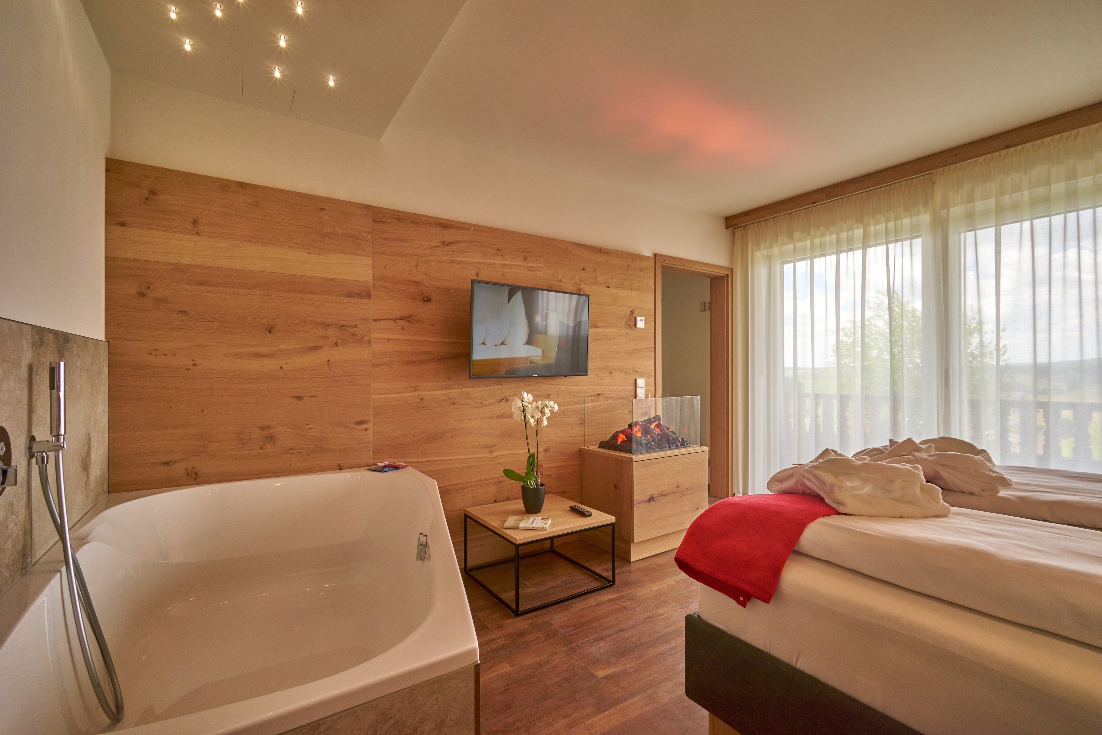 A modern hotel room with a wooden wall, a large bathtub, a bed with white linens, a small table with a vase of flowers, and a television mounted on the wall.