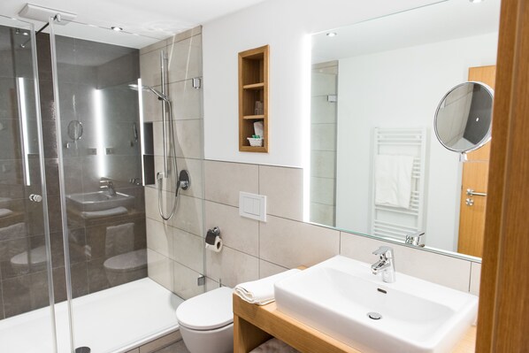 Un baño moderno con ducha envuelta en vidrio, lavamanos blanco y tocador de madera.