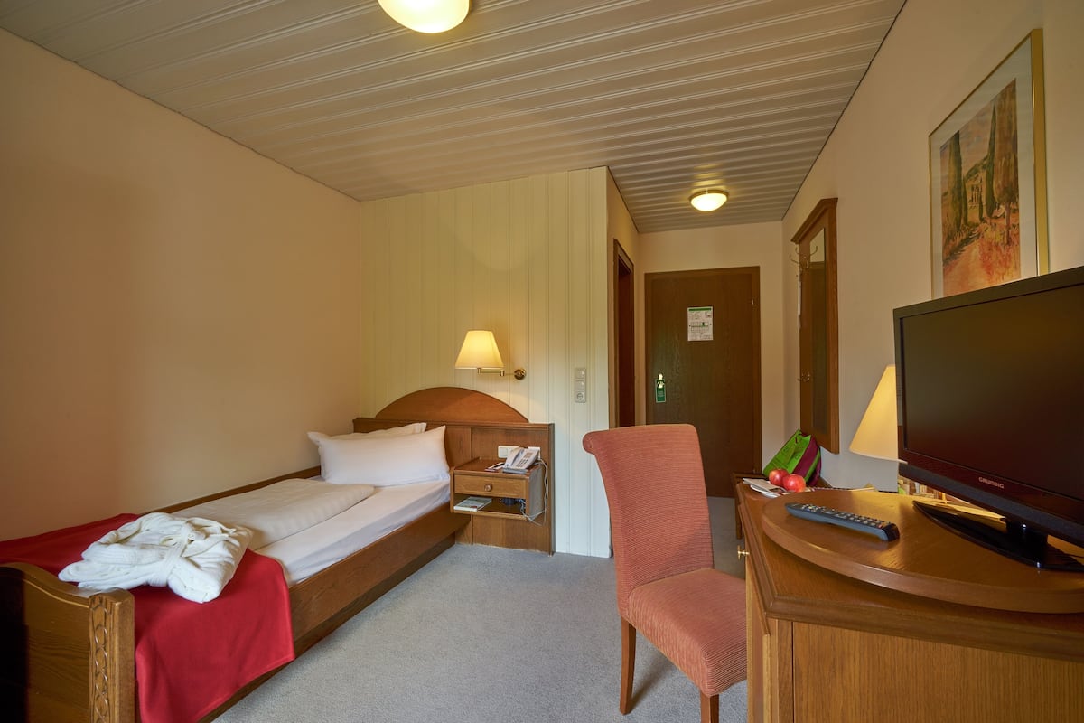 A hotel room with a single bed, a desk with a TV, a chair, a nightstand with a lamp, and a door to the bathroom.