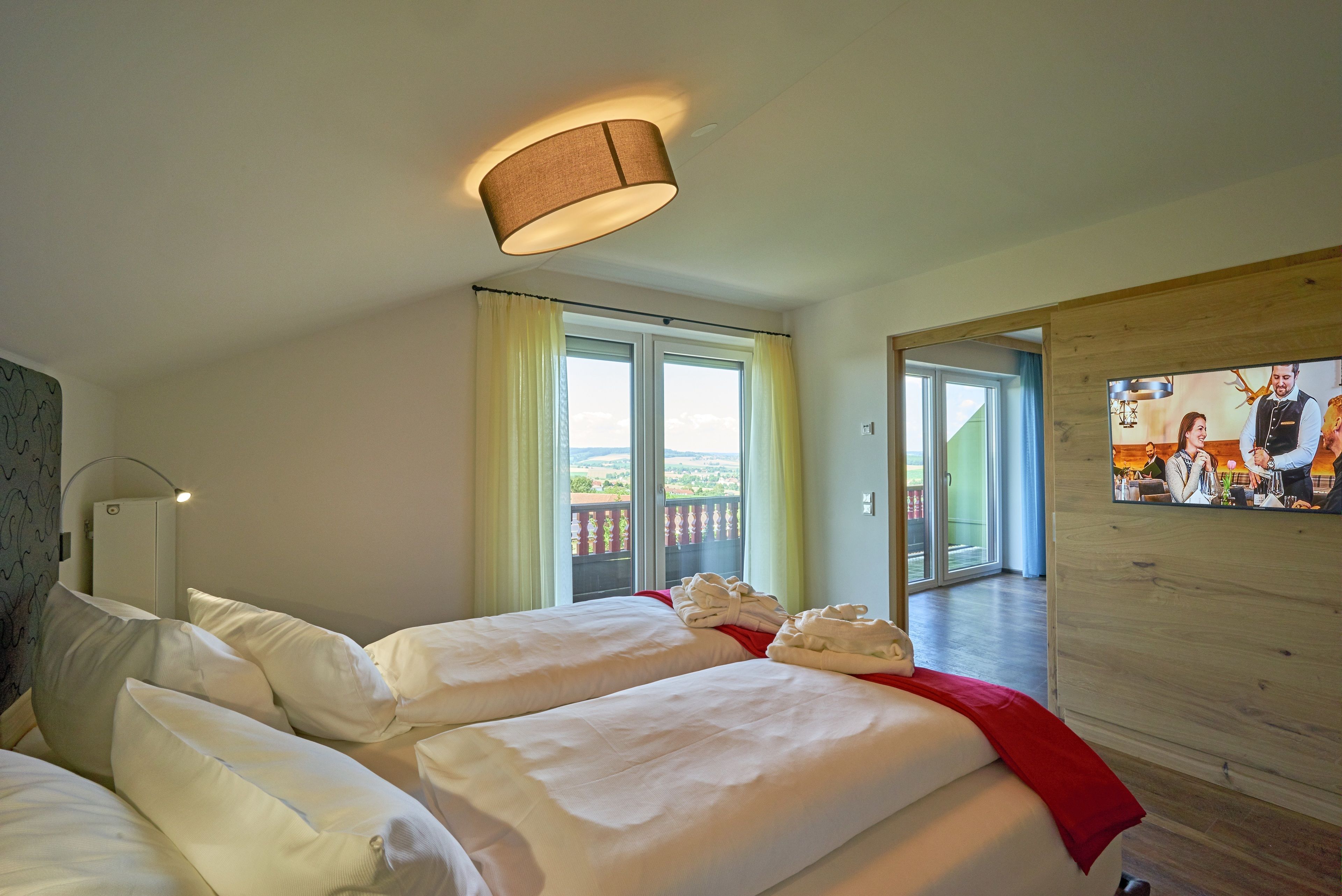 A hotel room with two beds, a large window with curtains, a ceiling lamp, and a television on the wall.
