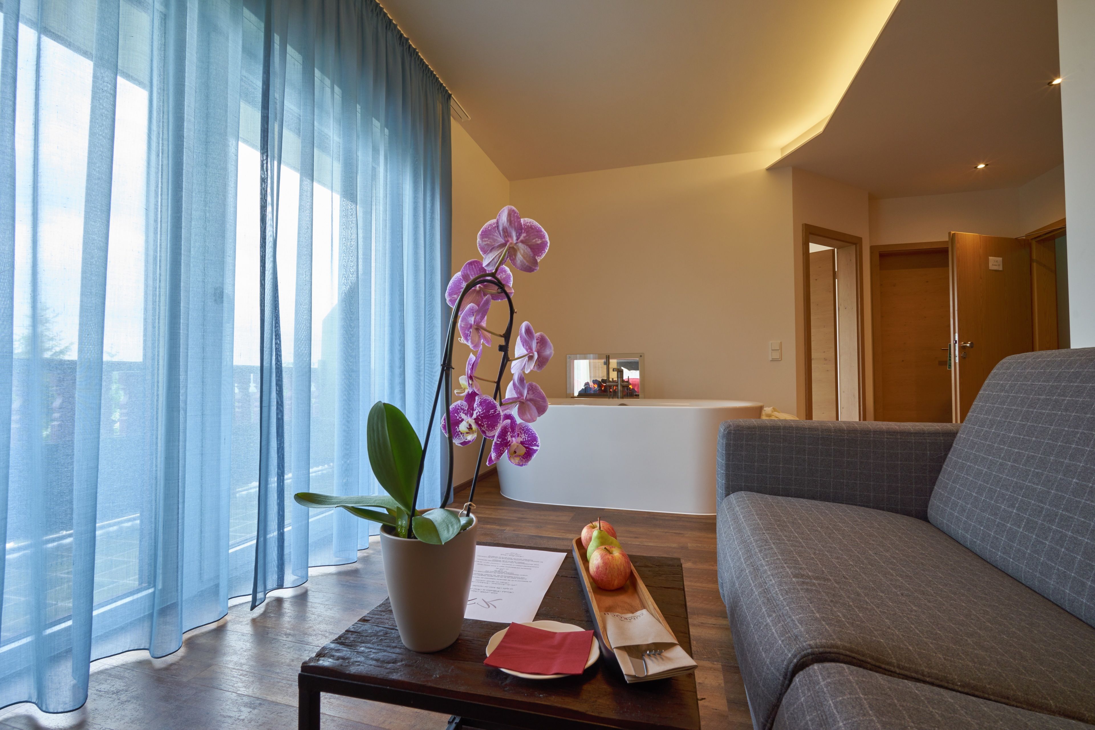 A modern living room with a sofa, a coffee table with a plant and apples, and a view of a reception area through sheer curtains.
