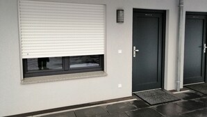 A modern building facade with a large window covered by horizontal blinds, a dark door with a handle, and a small vent above the door. - Landgasthof Waldrast (Voehrenbach)