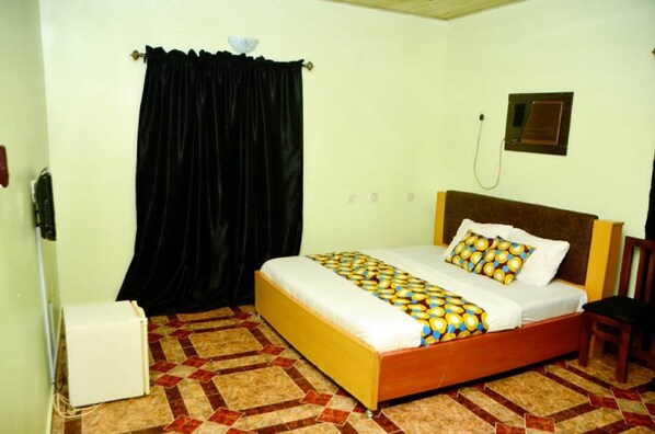Una cama individual con una colcha estampada, una silla, un calentador blanco pequeño y una cortina negra.